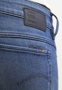 Tmavě modré džíny z denimového materiálu mají koženou nášivku s nápisem "RAW", s jemnými detailem šití a malou kovovou visačkou na kapse.