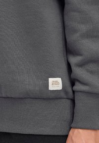 Grijze sweatshirt met ribgebreide manchetten en onderrand. Zachte stof met een klein wit label met "DBL END" in bruin. Close-up weergave van het kledingstuk.