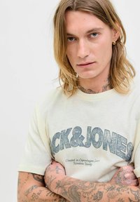 Grietos spalvos marškinėliai su "Jack & Jones" grafika struktūruotais mėlynais rašiniais. Artimo plano vaizdas pabrėžia tatuiruotes ir peties siūles.
