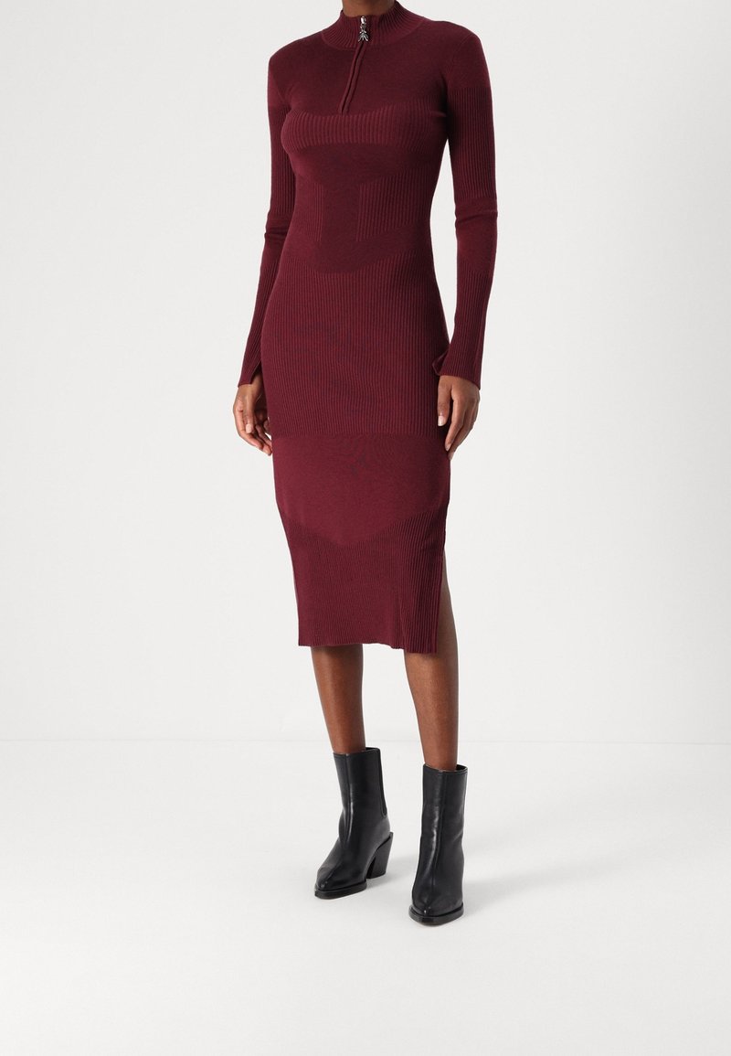 Femme portant une robe midi en maille côtelée bordeaux avec manches longues et col zippé, associée à des bottines noires à talons moyens.