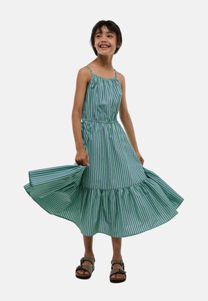 Robe rayée verte et blanche avec un bas évasé, des bretelles fines et une taille élastique. Portée avec des sandales marron.