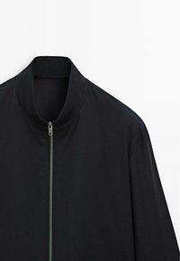 Black jacket with a silver front zipper and a short stand collar on a white background, showing the upper half and sleeves.