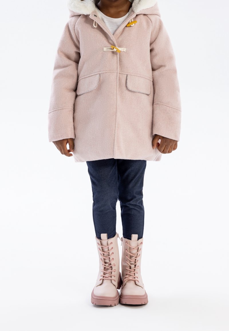 Enfant portant un manteau à capuche rose, un jean foncé et des bottines à lacets rose pâle, debout sur un fond blanc.