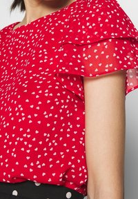Rød bluse med transparente ærmer, som har et hvidt hjerte mønster. Stof er let og struktureret, parret med en sort prikket talje.