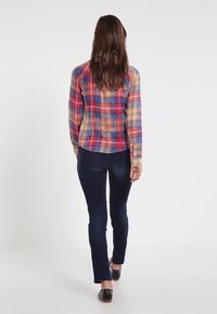 Kvinna med långt brunt hår som går bortåt, iförd en flerfärgad rutig skjorta, mörkblå jeans och svarta skor mot en enkel vit bakgrund.