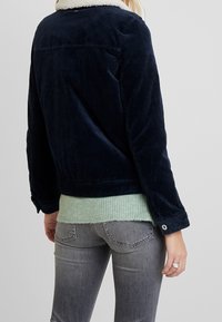 Marinblå kordjacka med en vit fleecekrage, med knappstängning och ribbade ärmmuddar, i kombination med en mintgrön stickad tröja under.