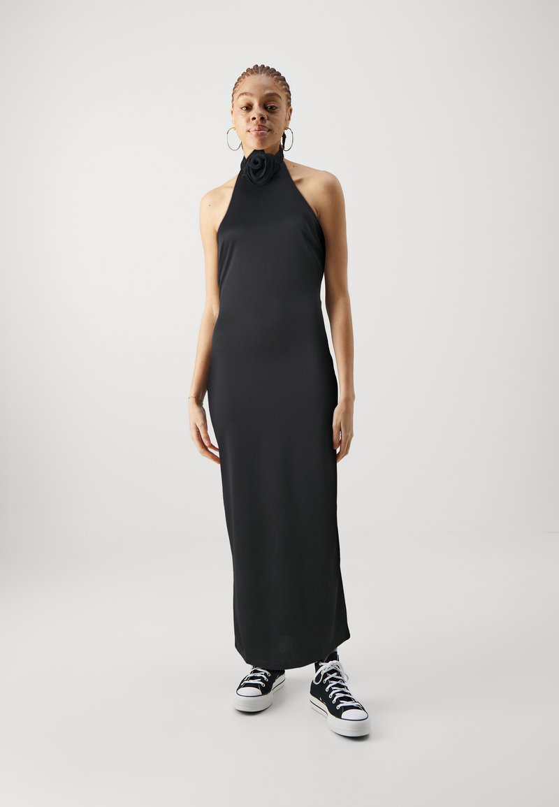 Črna halter obleka z cvetličnim naglasom okoli vratu, prilegajočega kroja, gladke teksture in dolge dolžine, v kombinaciji s črnimi supergami.