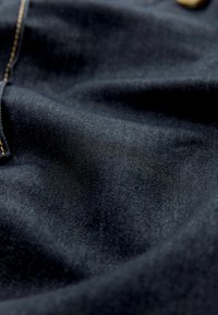 Donkerblauwe denimstof met een gladde textuur en zichtbaar weefsel. Heeft gouden stiknaden en een metalen knoopdetail.