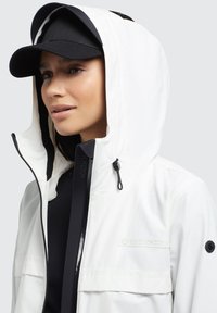 Veste imperméable blanche avec un col haut, des accents noirs et une fermeture éclair à l'avant. Elle est dotée d'une capuche ajustable et d'un logo sur la poitrine. Casquette noire portée.