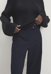 Tummansininen neulottu neule, jossa on leveät hihat, yhdistettynä räätälöityihin tummansinisiin housuihin, joissa on nappisulku ja etutaskut.