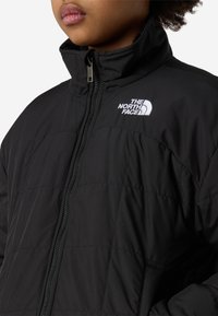 Μαύρο μπουφάν puffer με ψηλό γιακά, εμπρόσθιο φερμουάρ και λευκό λογότυπο στο αριστερό στήθος. Το υλικό είναι λείο με κεντητό σχέδιο.