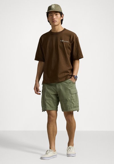 Rjav bombažni t-shirt z logotipom, zeleni cargo kratke hlače z stranskimi žepi in bež superge, na vrhu pa lahka zelena kapa.