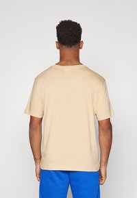 Beige pamut póló rövid ujjakkal és kerek nyakkivágással, kék rövidnadrággal viselve, egyszerű, díszítetlen designt mutatva.