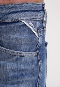 Blå denimjeans med medelhög midja, med en sydd ficka och en vit etikett med "REPLAY" på baksidan av midjebandet.