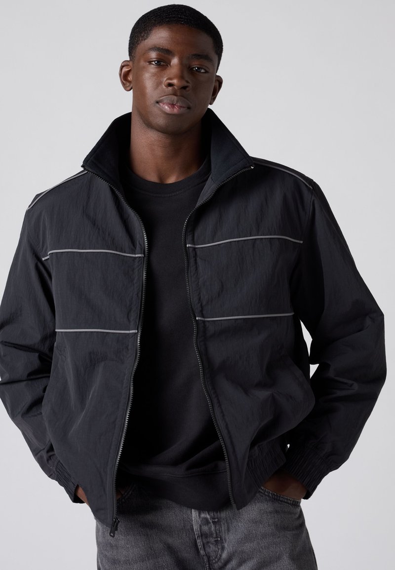 Jonge man draagt een zwarte jas met ritssluiting en reflecterende horizontale strepen, een zwart shirt en grijze spijkerbroek, handen in zijn zakken, neutrale achtergrond.