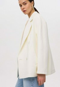 Crème op maat gemaakte blazer, dubbelgebreid ontwerp, gestructureerde schouders en zijzakken. Draag over een lichtgekleurde top en jeans.