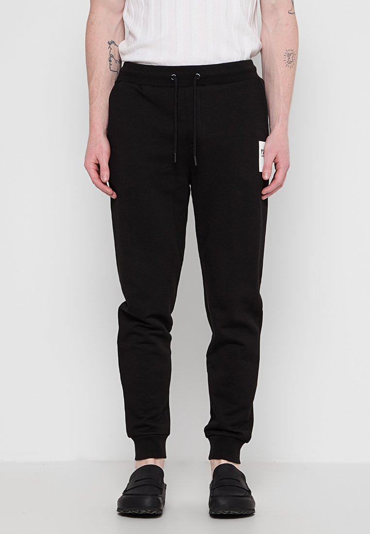 Schwarze Sweatpants aus weichem Material, schmale Passform, elastischer Bund mit Kordelzug, tapered Beinen, mit einem kleinen weißen Etikett an der Seite.