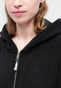 Svart zip-up hettegenser med teksturert overflate, hette og sølvfarget hardware. Har en subtil logo brodert på brystet og dekorativ glidelåsdrager.