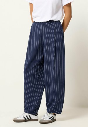 Osoba ubrana w szerokie spodnie w granatowym kolorze w prążki, białą koszulkę i białe trampki z czarnymi paskami, stojąca z rękami za plecami.