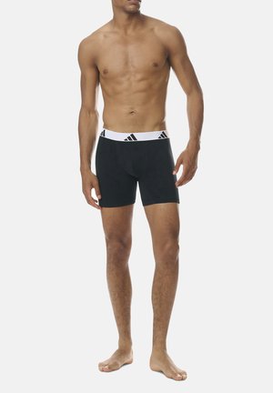 Black boxer shorts made of soft fabric with a white elastic waistband featuring black Adidas logos. Fitted design that extends to mid-thigh.