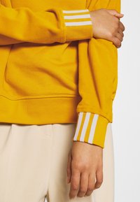 Gul fleece-pullover med ribbade ärmslut i vitt. Mjuk textur, med en avslappnad passform och en enfärgad design utan mönster.