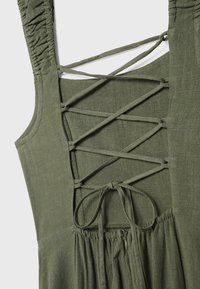 Vestido verde oliva con tejido texturizado, tirantes anchos y una espalda con lazada cruzada y detalle de lazo. Cintura fruncida con cremallera.