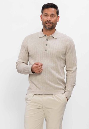 Man draagt een beige geribd poloshirt met lange mouwen en lichtbeige broek, staat met één hand in de zak en de andere hand omhoog bij de borst.