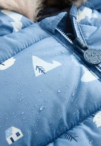 Water-resistant blue padded jacket featuring white mountain and house patterns. Close-up view with droplets on the fabric. Textured material.