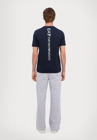 Man i marinblå t-shirt med vertikalt vitt "EA7 Emporio Armani"-logotyp på ryggen, ljusgråa sweatpants, står mot vit bakgrund.