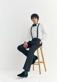 Formal outfit featuring a white shirt, black bow tie, dark trousers, green velvet shoes, and black suspenders, seated on a wooden stool.