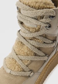 Bottine beige avec doublure en laine douce, suède texturé et lacets épais. Comprend des œillets métalliques et une semelle en caoutchouc rainurée.