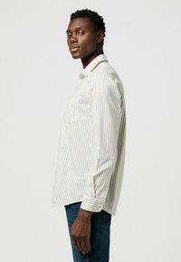 Camicia a maniche lunghe a righe di colore beige chiaro con sottili strisce marroni, dotata di una tasca sul petto e polsini con bottoni, abbinata a jeans blu scuro.