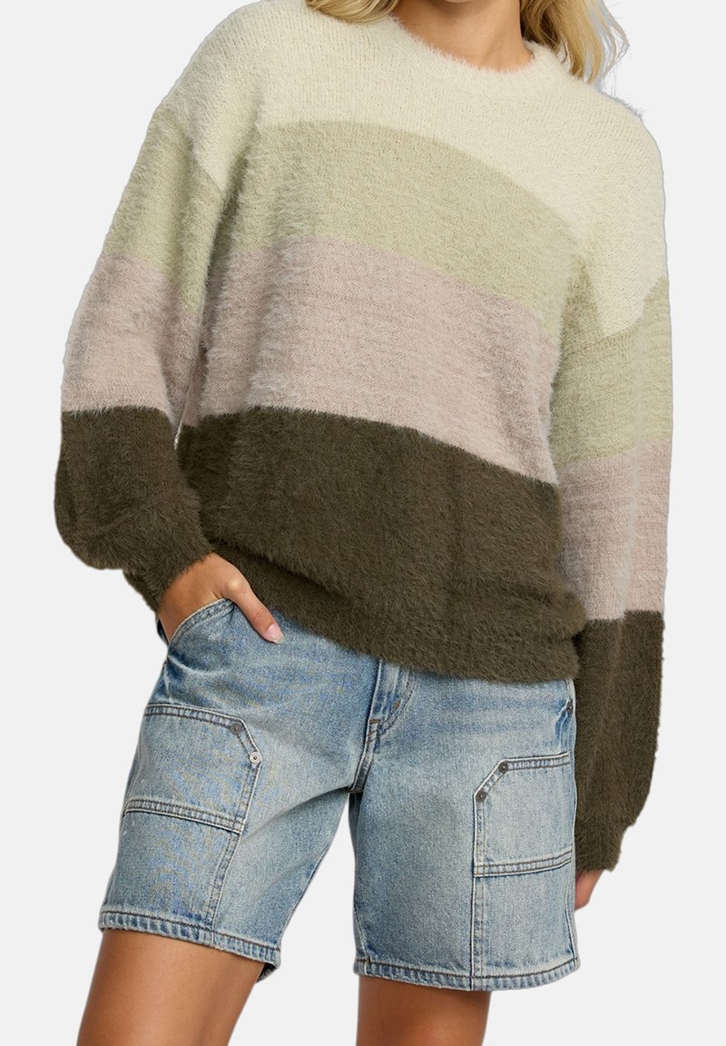 Femme portant un pull à blocs de couleur en matière duveteuse beige, rose et vert olive, avec un short cargo en denim bleu clair, main dans la poche.