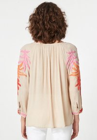 Blusa beige a maniche lunghe con ricami floreali colorati in rosa e arancione sulle maniche, caratterizzata da una schiena arricciata e una vestibilità rilassata.