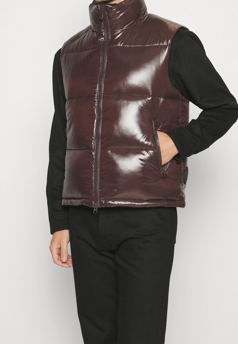 Braune glänzende Pufferweste mit hohem Kragen, Frontreißverschluss und zwei seitlichen Taschen. Getragen über einem schwarzen Langarmshirt.