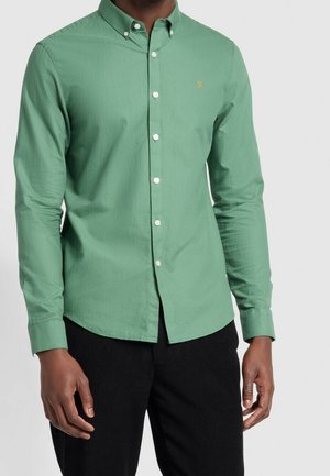 Man draagt een aansluitend groen overhemd met witte knopen en een klein geel geborduurd logo op de borst, gecombineerd met een zwarte broek.