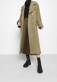 Lång, olivgrøn uldblandingsfrakke med dobbeltbrystet design, brede reverser og guldknapper, parret med en sort strikket top og plisseret nederdel.