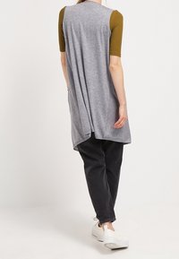 Túnica larga gris con un corte holgado, que presenta mangas cortas de color verde oliva, diseño suelto en la parte trasera y bolsillos laterales. Combinada con pantalones negros y zapatillas blancas.