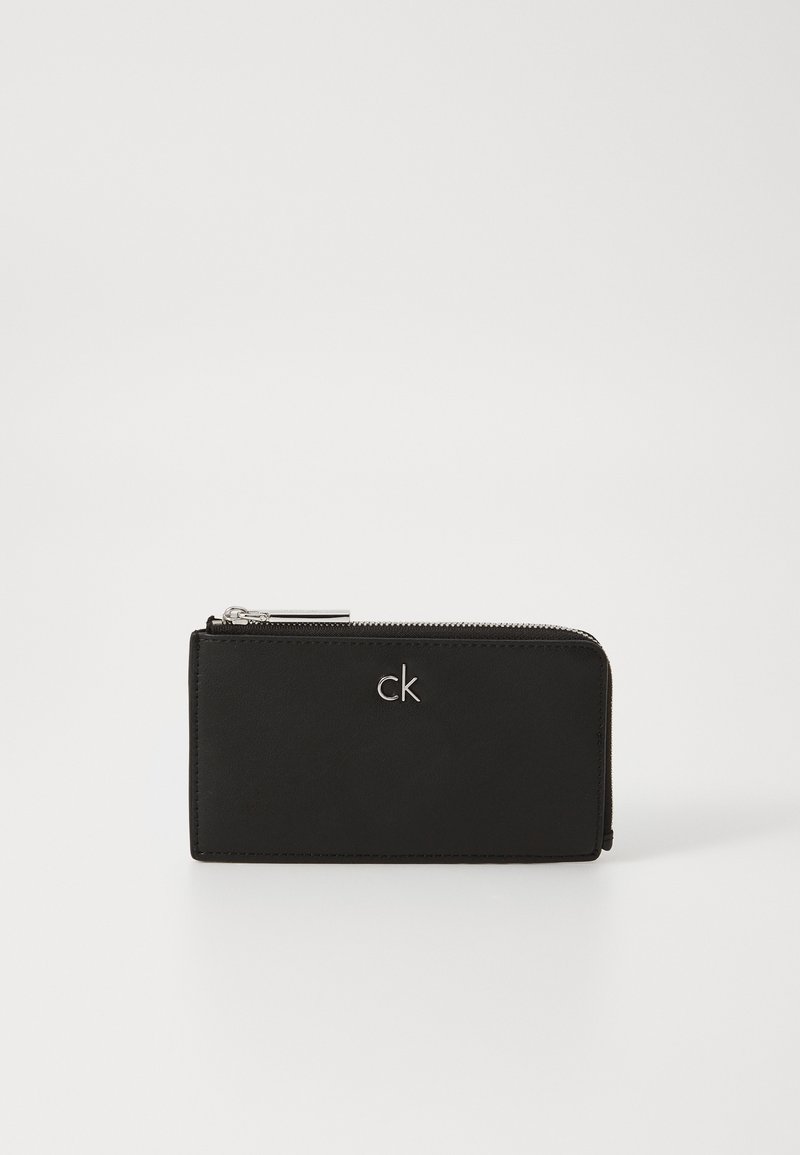 Cartera de cuero negro con textura suave, que presenta un zipper plateado y un sutil logo 'ck' en relieve en el frente.