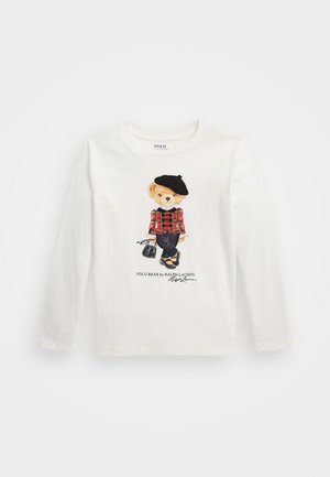 Chemise blanche à manches longues avec un ours imprimé portant un béret noir, une veste à carreaux rouge, un pantalon noir et tenant un sac à main.