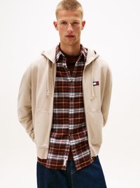Felpa beige con cappuccio e tasche frontali, indossata sopra una camicia a quadri marrone e blu. Il modello indossa jeans in denim blu scuro.
