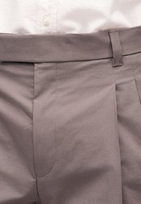 Κοντινή λήψη στη μέση παντελονιού τσόπ σε αποχρώσεις του ταουπ με θηλιά για ζώνη, φερμουάρ και μέρος μιας κρεμ πουκαμίσας που έχει μπει μέσα.