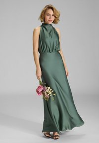 Langes grünes Satin-Kleid mit hohem Ausschnitt, geraffter Taille und fließendem Rock. Das Model hält einen Strauß pinkfarbener Blumen. Schwarze Sandalen runden den Look ab.