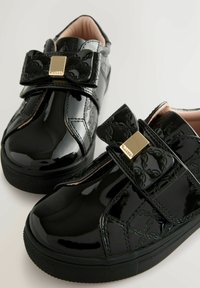 Scarpe in pelle verniciata nera con finitura lucida, dotate di un grande dettaglio a fiocco, cinturino in Velcro e dettagli testurizzati sulla parte superiore.