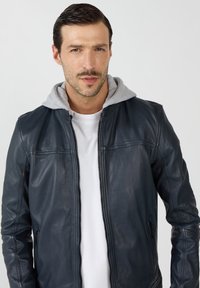 Marineblaues Lederjacke mit grauem Kapuzenfutter, Frontreißverschluss, Seitentaschen und glatter Textur, kombiniert mit einem weißen T-Shirt darunter.