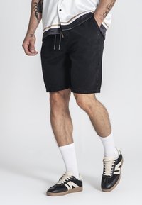Shorts en denim noirs avec une coupe décontractée, portés avec des chaussettes blanches jusqu'à la cheville et des baskets noires avec des accents blancs et beige.