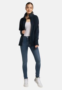 Marineblaues Zip-Jackett mit hohem Kragen, gerippten Bündchen und Logo-Detail, kombiniert mit hellblauen Skinny-Jeans und weißen Sneakern.