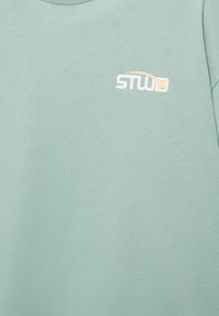 Material verde deschis cu un mic logo "STWL" alb și bej imprimat în partea stângă sus.