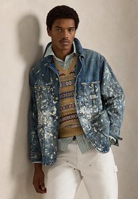 Νεαρός άνδρας που φοράει ένα τζιν μπουφάν με πιτσιλιές από μπογιά πάνω από ένα πουλόβερ με σχέδια και μια ριγέ πουκάμισο, στέκεται μπροστά σε ουδέτερο τοίχο.