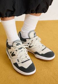Füße in weißen und schwarzen New Balance 550 Sneakers mit weißen Socken auf einem gelben Teppich, kombiniert mit schwarzen Hosen.